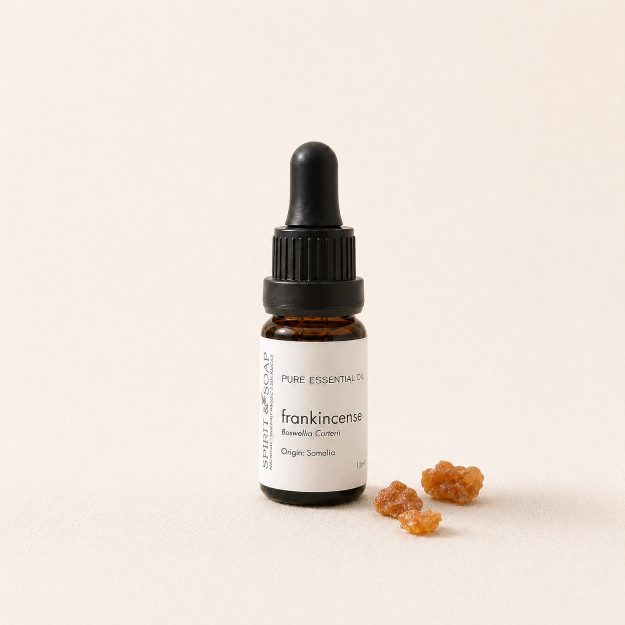 A small amber glass dropper bottle labeled frankincense essential oil, standing on a warm off-white background with a few amber resin pieces beside it. frankincense