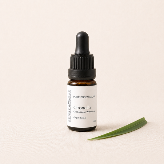 A small amber glass dropper bottle labeled 'PURE ESSENTIAL OIL citronella' with a black cap, standing on a light beige textured surface next to a green leaf.