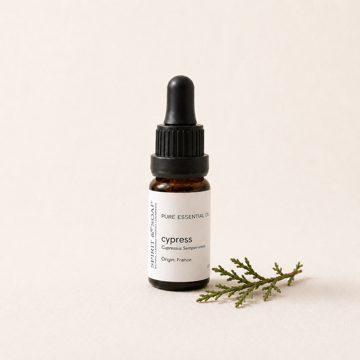 A small amber glass dropper bottle labeled PURE ESSENTIAL OILS cypress, with a black dropper cap and a white label, photographed on a warm off-white background with a small green sprig beside it.