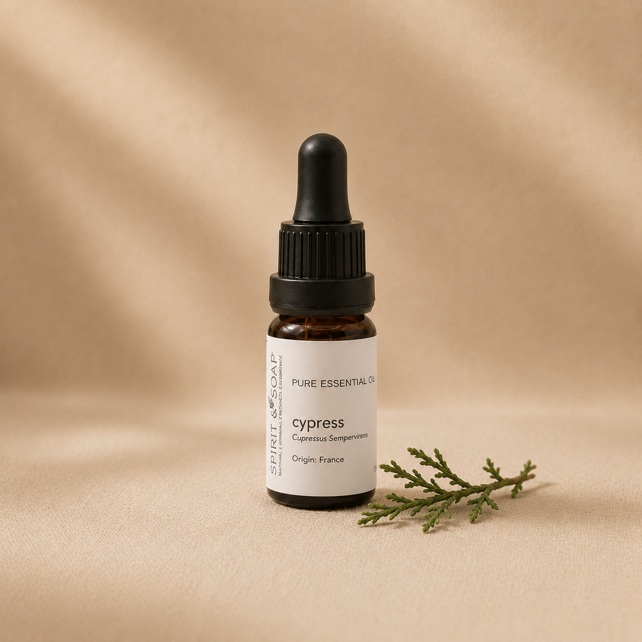 A small amber glass dropper bottle labeled 'cypress' with a black dropper cap, set on a warm beige textured surface with soft shadows and a small green sprig beside it.