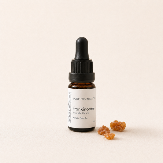 A small amber glass dropper bottle labeled frankincense essential oil, standing on a warm off-white background with a few amber resin pieces beside it. frankincense