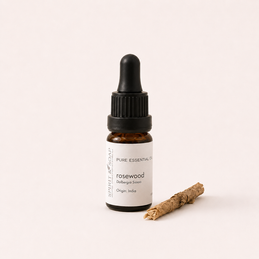 A small amber glass dropper bottle labeled 'rosewood' with a black cap and white label, standing on a light beige background with a small rolled wood stick beside it.